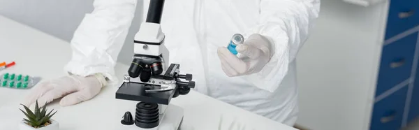 Cropped view of scientist in protective uniform holding vaccine near microscope and pills, banner — Stock Photo