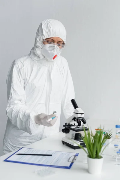 Scientist in hazmat suit looking at vaccine near clipboard and microscope in laboratory — Stock Photo