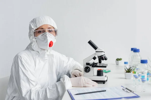 Вчений у захисній масці та окулярах дивиться на камеру поблизу мікроскопа, вакцини та буфера — стокове фото
