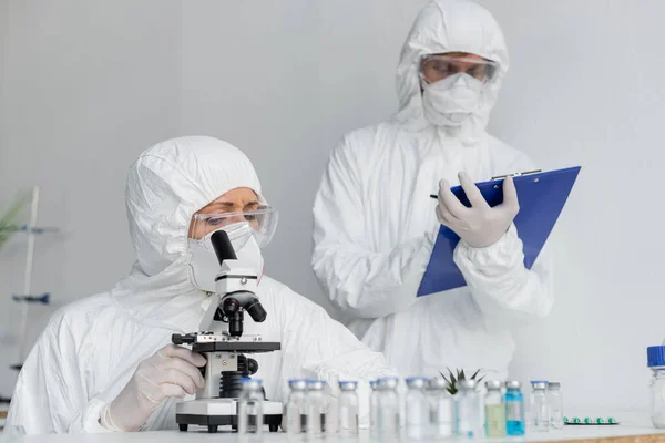 Вчений в захисній формі з використанням мікроскопа біля колеги і вакцин на розмитому передньому плані — стокове фото