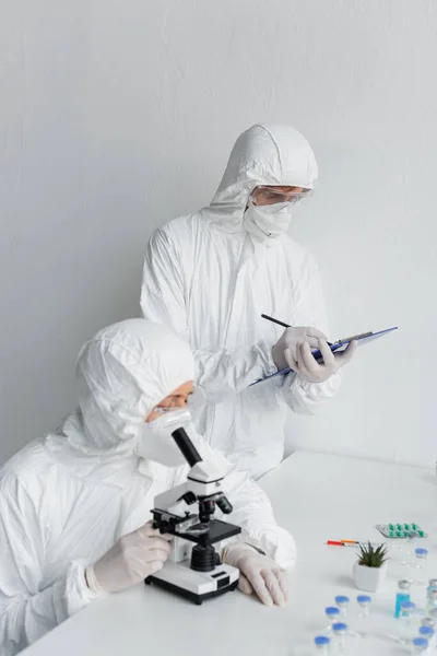 Вчений у костюмі ризику, який вивчає вакцину поблизу колеги, пишучи на кишені в лабораторії — стокове фото
