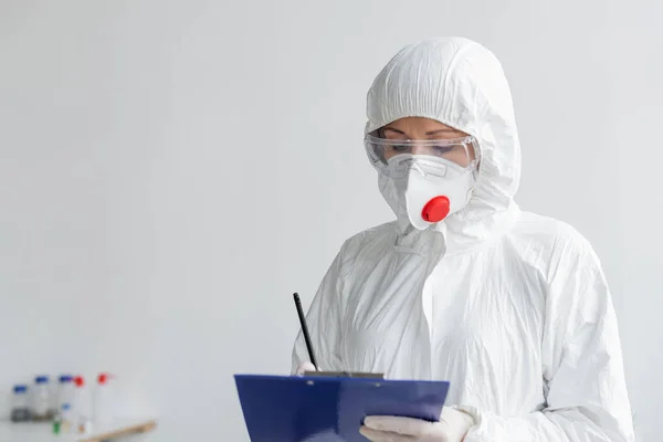 Вчений в медичній масці і захисний костюм, що пише на кишені на розмитому передньому плані — стокове фото