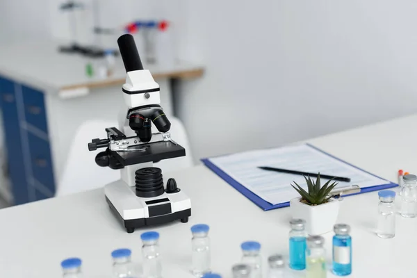 Microscope near vaccines and clipboard on blurred background in laboratory — Stock Photo