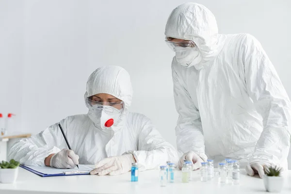 Вчений пише на кишені біля колеги і вакцини на розмитому фоні — стокове фото