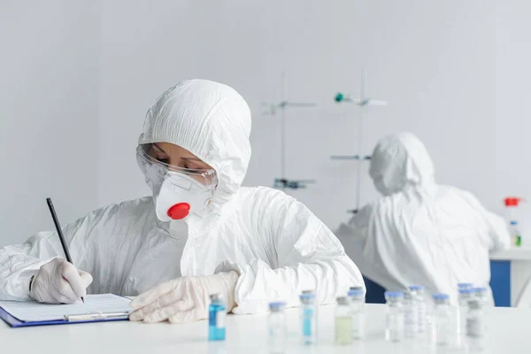 Scientist in protective uniform writing on clipboard near vaccines and colleague on blurred background — Stock Photo
