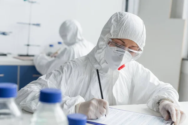 Scientist writing on clipboard near colleague on blurred background in laboratory — Stock Photo