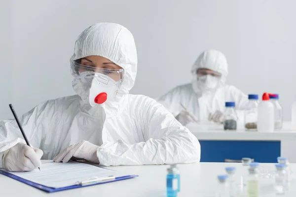Scientist in protective suit and latex gloves writing on clipboard near vaccines and colleague — Stock Photo