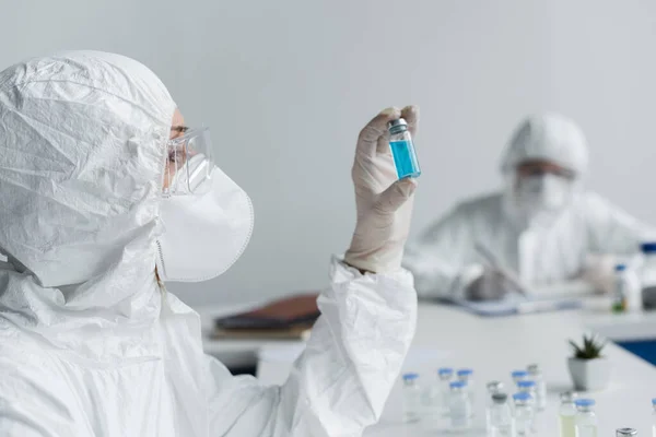 Scientist looking at jar with vaccine near colleague on blurred background — Stock Photo