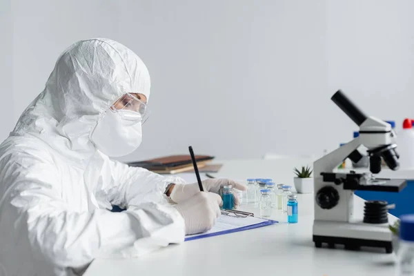 Scientist in protective uniform writing on clipboard near microscope and vaccines — Stock Photo