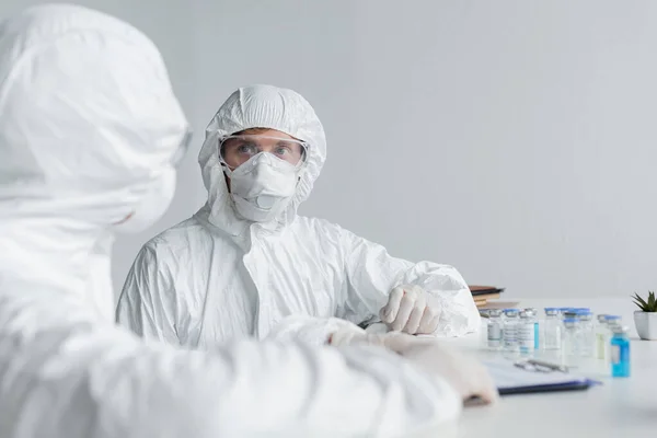 Scientist in hazmat suit looking at colleague near clipboard and vaccines — Stock Photo