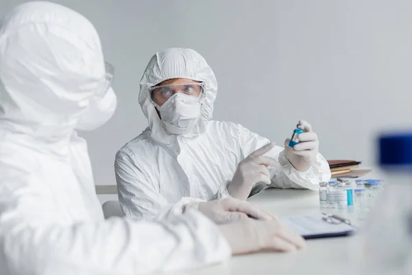Scientist in protective suit pointing at vaccine near colleague on blurred foreground in laboratory — Stock Photo