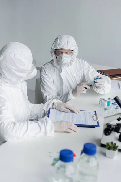 Scientists working with vaccines and clipboard near microscope on blurred foreground — Stock Photo