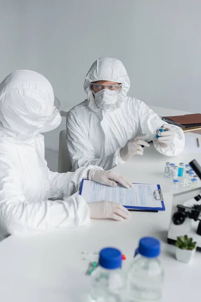 Científicos trabajando con vacunas y portapapeles cerca del microscopio en primer plano borroso - foto de stock