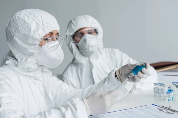 Wissenschaftler nimmt Impfstoff in Spritze neben Klemmbrett auf und Kollege auf verschwommenem Hintergrund — Stockfoto