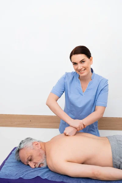 Massagista sorrindo para a câmera ao fazer massagem nas costas para o homem no centro de reabilitação — Fotografia de Stock