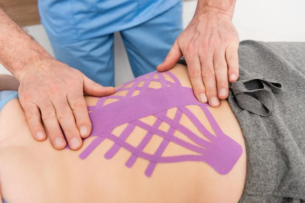 Vista parcial del rehabilitólogo haciendo el procedimiento de kinesiotaping en la mujer en la clínica - foto de stock