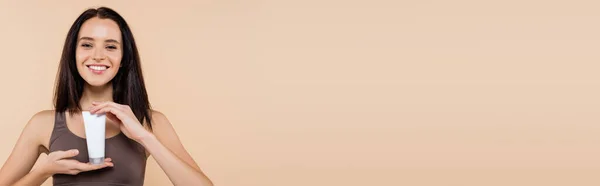 Jeune femme souriant à la caméra tout en tenant la crème cosmétique isolée sur beige, bannière — Photo de stock