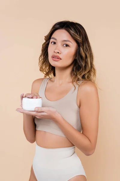 Hübsche asiatische Frau hält kosmetische Creme in der Hand, während sie isoliert auf beige in die Kamera schaut — Stockfoto