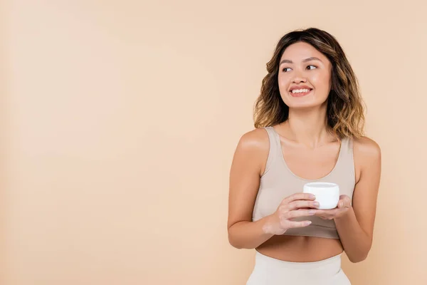 Sonriendo mujer asiática mirando hacia otro lado mientras sostiene crema cosmética aislado en beige - foto de stock