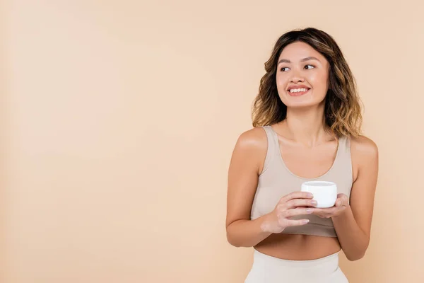 Smiling asian woman looking away while holding cosmetic cream isolated on beige — Stock Photo