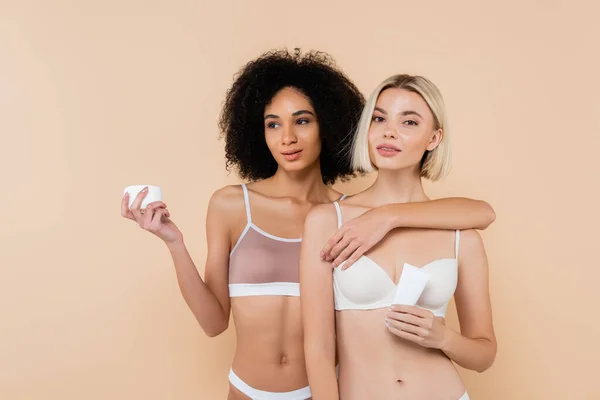 Slim multiethnic women in underwear holding cosmetic cream isolated on beige — Stock Photo