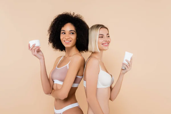 Sonriente interracial las mujeres celebración cosmética crema mientras de pie espalda con espalda aislado en beige - foto de stock