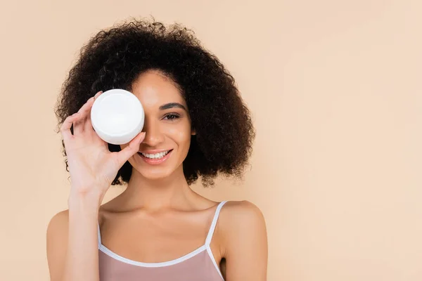 Cheerful african american woman covering eye with container of cosmetic cream isolated on beige — Stock Photo