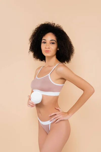 Mujer afroamericana sosteniendo crema cosmética mientras posa con la mano en la cadera aislada en beige - foto de stock