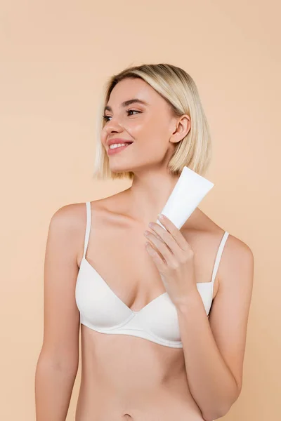 Mujer alegre en sujetador blanco mirando hacia otro lado mientras sostiene crema cosmética aislada en beige - foto de stock
