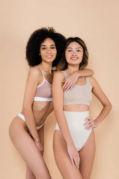 African american woman hugging asian friend while posing in underwear isolated on beige — Stock Photo