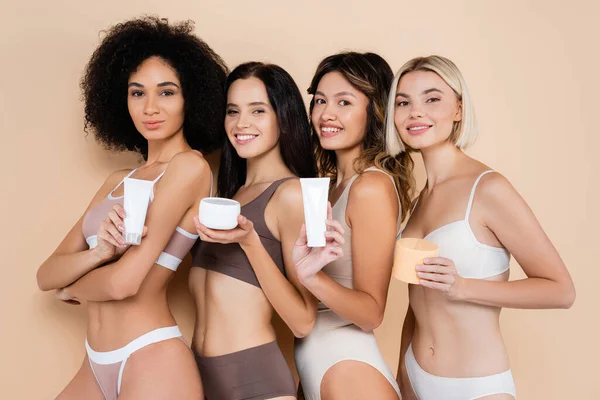 Sexy multiethnique femmes tenant crème pour le corps tout en souriant à la caméra sur beige — Photo de stock
