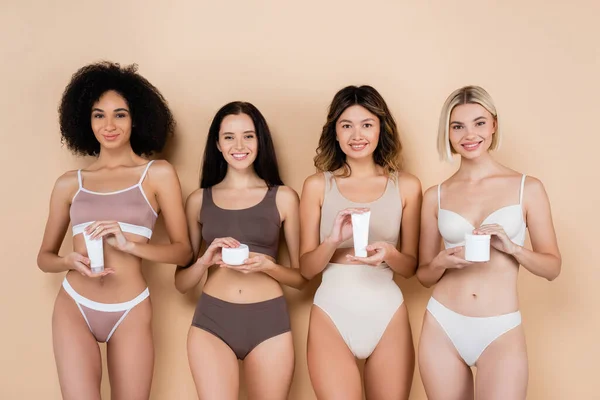 Vorderseite interrassische Frauen mit perfekten Körpern, die kosmetische Creme auf beige halten — Stockfoto