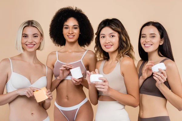 Fröhliche multikulturelle Frauen in Unterwäsche posieren mit kosmetischer Creme isoliert auf beige — Stockfoto
