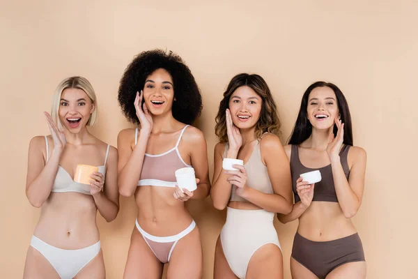 Amazed interracial women in underwear applying face cream on beige — Stock Photo