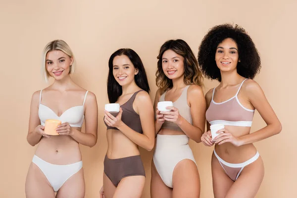 Sexy interracial femmes en sous-vêtements tenant crème pour le corps sur beige — Photo de stock