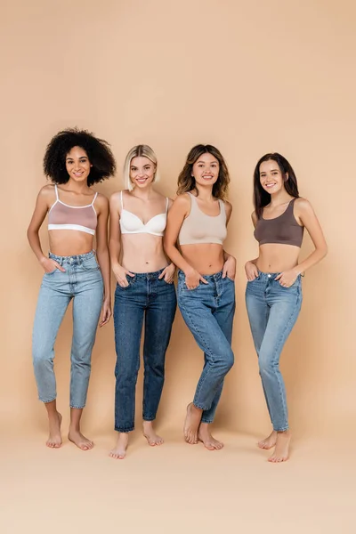 Повнометражний вид сексуальних міжрасових жінок позує в джинсах і бюстгальтерах на бежевому — стокове фото
