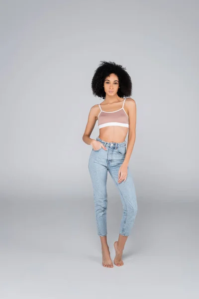 Full length view of slim african american woman posing in bra and jeans on grey — Stock Photo