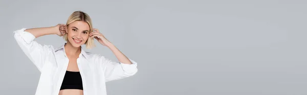 Donna bionda in camicia bianca sorridente alla macchina fotografica mentre fissa i capelli isolati sul grigio, banner — Foto stock