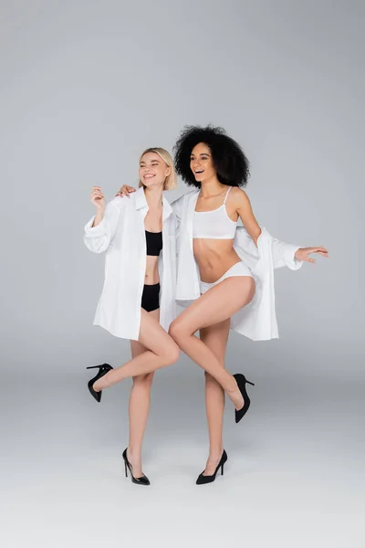 Cheerful interracial women in underwear and white shirts posing on heels on grey — Stock Photo