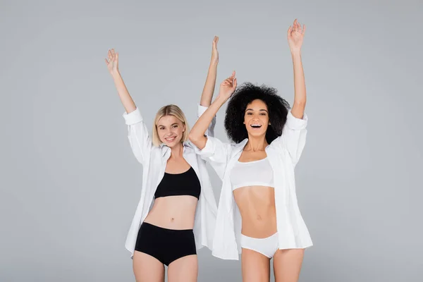 Excited multiethnic women in underwear and white shirts posing with raised hands isolated on grey — Stock Photo