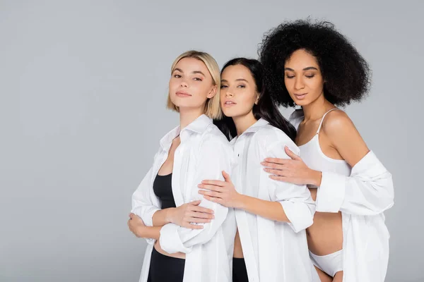 Jóvenes mujeres interracial en ropa interior y camisas blancas mirando a la cámara aislada en gris - foto de stock