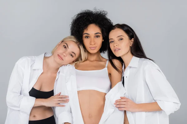 Mujeres bonitas en camisas blancas y ropa interior apoyada en amigo afroamericano aislado en gris - foto de stock