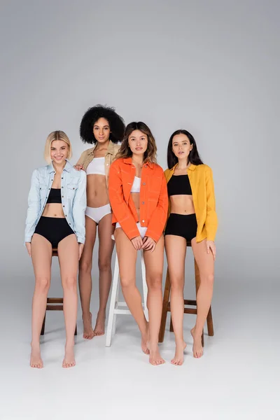 Sexy interracial femmes en sous-vêtements et chemises regardant caméra près de hauts tabourets sur gris — Photo de stock