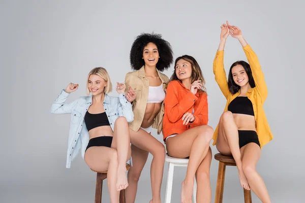 Happy african american women pointing with finger near interracial friends posing on high stools on grey — Stock Photo