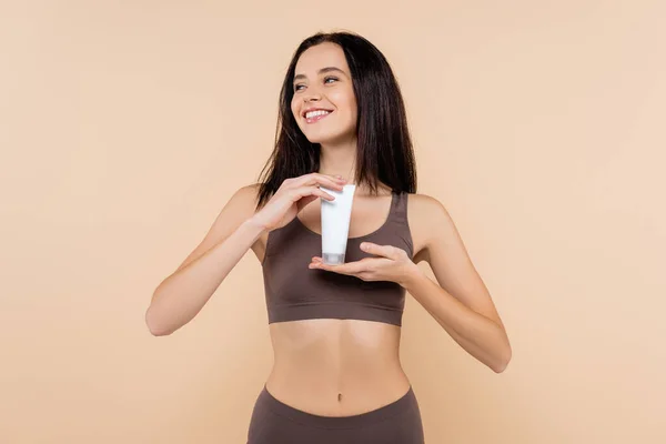 Sorrindo mulher olhando embora ao mostrar creme cosmético isolado no bege — Fotografia de Stock