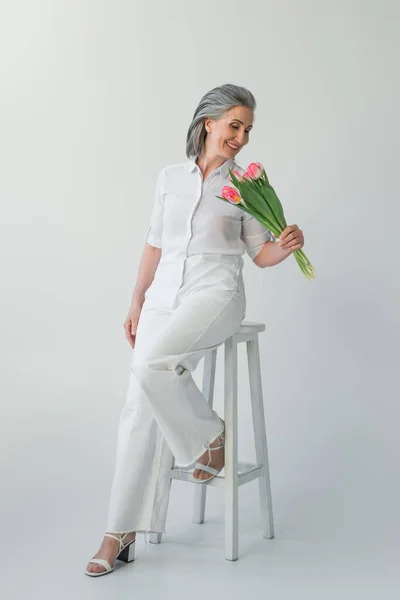 Mujer madura sonriente mirando tulipanes cerca de la silla sobre fondo gris - foto de stock