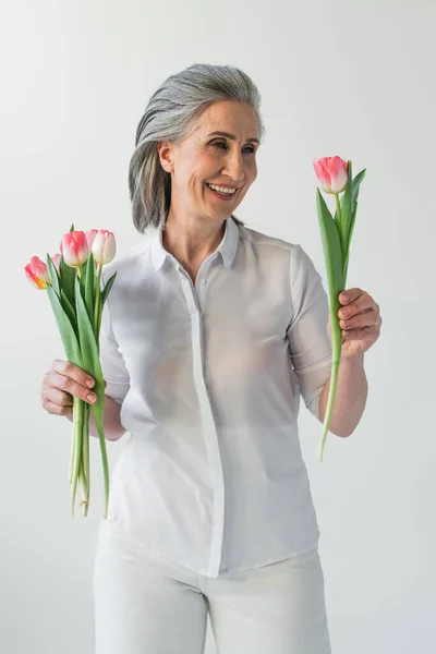 Весела зріла жінка дивиться на тюльпан ізольовано на сірому — стокове фото
