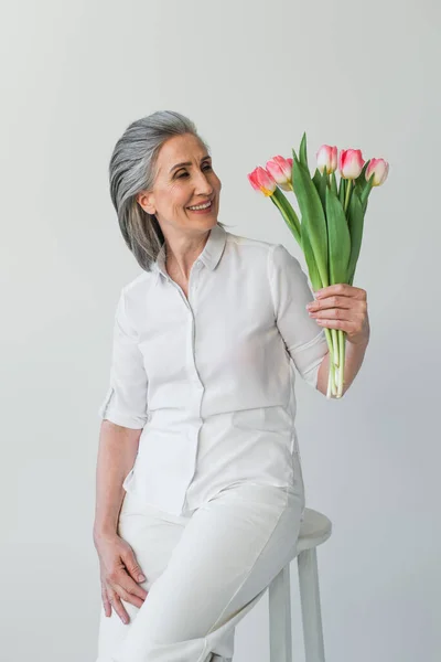 Smiling grey haired woman looking at bouquet of tulips isolated on grey — Stock Photo