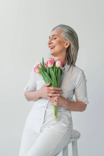 Ältere Frau mit geschlossenen Augen lächelt, während sie Tulpen isoliert auf grau hält — Stockfoto