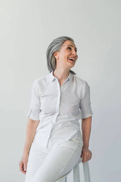 Grauhaarige Frau im weißen Hemd schaut weg auf Stuhl isoliert auf grau — Stockfoto